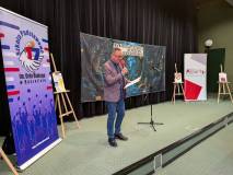 Mężczyzna w okularach, ubrany w fioletową marynarkę, niebieską koszulę i jeansy, stoi na scenie i pr