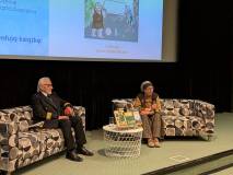 Zbliżenie na dwoje prelegentów siedzących na czarno-białych fotelach na scenie. Po lewej stronie męż