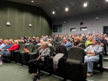 Widok z boku na wypełnioną salę widowiskową. Rzędy ciemnozielonych foteli zajęte są przez osoby w ró