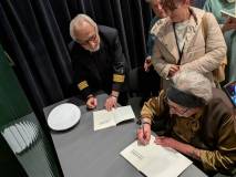 Anna Bojar - Fijałkowska siedzi przy szarym stoliku i wpisuje dedykację do otwartej książki. Obok ni