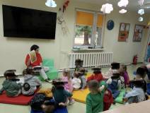 Dzieci siedzą na kolorowych materacykach a pani bibliotekarka pokazuje im kolorowe plansze.