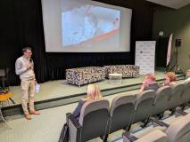 Mężczyzna w okularach, jasnym swetrze i beżowych spodniach stoi na scenie i przemawia do mikrofonu p