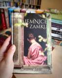 Dłoń trzyma książkę Tajemnice zamku autorstwa Lucinda Riley. Na okładce widać kobietę w różowej suki