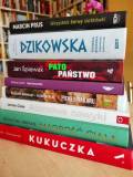 Stos książek różnych autorów ustawiony na stole. W tle rozmyty regał z książkami.