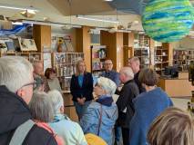 Zbliżenie na prelegentów i uczestników w bibliotece. Centralną postacią jest kobieta w ciemnej maryn