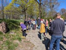 Grupa kilkunastu osób stoi na brukowanej alejce w parku. Większość patrzy w stronę dużego kamienia p