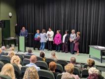 zeroki plan na scenę z ciemną kotarą w tle.

Postacie: Na scenie stoi grupa ośmiu kobiet w rzędzie