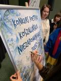 Zbliżenie na dłoń podpisującą czarnym markerem biało-niebieski plakat z napisem „KONKURS POETYCKI KO