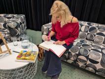 Justyna Sobolewska w czerwonej bluzce siedzi na kanapie i podpisuje egzemplarz swojej książki. Obok,