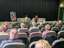 Zdjęcie wykonane z perspektywy widzów w sali kinowej. Widać rzędy foteli z publicznością oraz scenę,