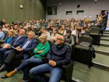Widok z boku na wypełnioną salę kinową lub audytoryjną. W pierwszym rzędzie siedzi pięcioro dorosłyc