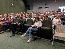 Widok z boku na widownię sali kinowej wypełnioną publicznością, składającą się głównie z osób w śred