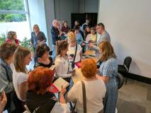 Grupa ludzi zgromadzona wokół Joanny Brodzik w jasnym korytarzu budynku. Aktorka, ubrana w błękitną 