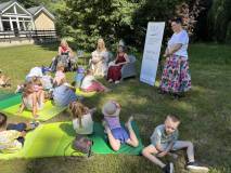 Dzieci siedzą i leżą na zielonych matach rozłożonych na trawie podczas plenerowego spotkania czyteln