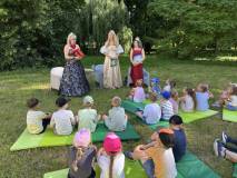 Dzieci siedzą i leżą na zielonych matach rozłożonych na trawie podczas plenerowego spotkania czyteln