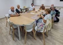 Siedem osób spotyka się przy dużym, owalnym stole. Uczestnicy trzymają książki i rozmawiają przy kaw