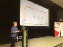 Prelegentka w niebieskiej koszuli stoi przy ambonce konferencyjnej, obsługując laptopa. Na ekranie z