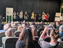 Na scenie przed czarną kurtyną stoi grupa osób ubranych w stroje z ozdobnymi frędzlami. Po prawej st