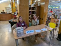 Dwie kobiety siedzą za drewnianym stołem w bibliotece. Jedna ma na sobie bordową bluzkę w kwiaty, dr