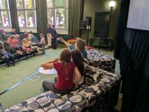 W sali bibliotecznej dzieci i dorośli siedzą na fotelach i kanapach. Mężczyzna w dżinsowej koszuli f