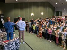 Adam Mirek i prowadzący spotkanie stoją przodem do publiczności w sali bibliotecznej. Dzieci i młodz