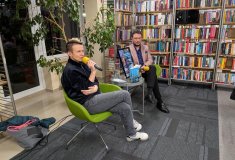 Kobieta siedząca na zielonym fotelu wśród bibliotecznych regałów trzyma w dłoni żółty mikrofon i wyp