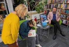 Spotkanie autorskie w bibliotece. Na pierwszym planie kobieta w żółtym swetrze obserwuje osobę siedz