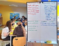 Widok na dzieci pracujące przy stolikach w bibliotece, obok dużej tablicy z odręcznymi notatkami. Ta
