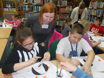 Dzieci siedzą przy stole i malują, a bibliotekarki wspierają ich prace