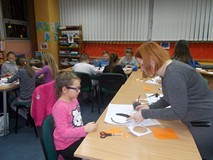 Dzieci siedzą przy stole i wykonują pracę plastyczną, a bibliotekarka pomaga dziewczynce malować pin