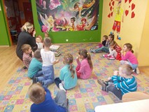 Dzieci siedzą na dywanie i naśladują bibliotekarkę, która pokazuje palcem na swój nos