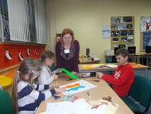 Dzieci siedzą przy stole i wycinają elementy, a bibliotekarka obserwuje ich pracę