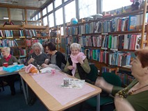 Panie siedzą przy stole i pokazują swoje szale robione na drutach