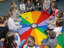 Dzieci z bibliotekarką siedzą na chuście animacyjnej i rzucają kostką