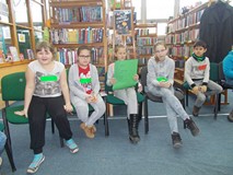 Dzieci siedzą na krzesłach, a jedna dziewczynka czyta odpowiedzi drużyny