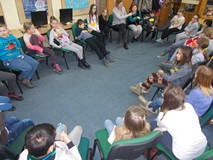 Dzieci siedzą w kole na krzesłach, a bibliotekarki pokazują im gesty do naśladowania