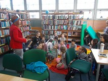 Bibliotekarka stoi przed dziećmi siedzącymi na chuście animacyjnej mając na głowie kartkę z ilustrac