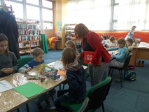 Bibliotekarka stoi przy dzieciach siedzących przy stole pomagając im w trakcie pracy plastycznej