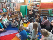 Dzieci siedzą na chuście animacyjnej, a bibliotekarka prezentuje książkę detektywistyczną