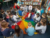 Dzieci siedzą na chuście animacyjnej i patrzą na bibliotekarkę, trzymającą książkę detektywistyczną