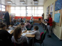 Dzieci siedzą przy stołach, a bibliotekarka objaśnia im zabawę