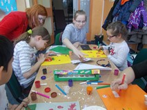 Dzieci siedzą przy stole, a bibliotekarka pomaga dziewczynce w pracy plastycznej