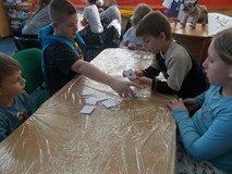 Dzieci siedzą przy stole i odkrywają kartki ze swoimi odciskami palców