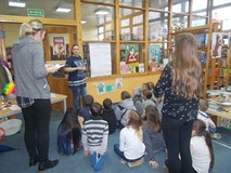 Dzieci siedza na dywanie i obserwują dziewczynę sstojącą przy brystolu, a bibliotekarka czyta hasła 