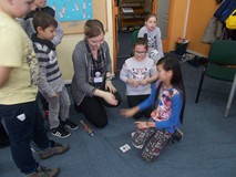 Dzieci wraz z bibliotekarką siedzą, a dziewczynka pokazuje karty do gry