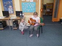 Dziewczynka siedzi z gitarą, obok dziewczynka trzyma tekst