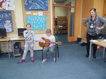 Dziewczynka siedzi na krześle z gitarą, obok stoi bibliotekarka i dziewczynka