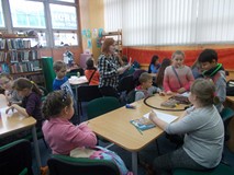 Dzieci siedzą przy stole, a bibliotekarka wyjaśnia zasady następnego zadania