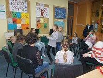 Dwie dziewczynki stoją w środku koła. Pozostałe dzieci wraz z bibliotekarką siedzą na krzesłach w ko