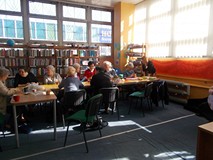 Panie siedzą przy stołach a bibliotekarka ogląda ich rękodzieła